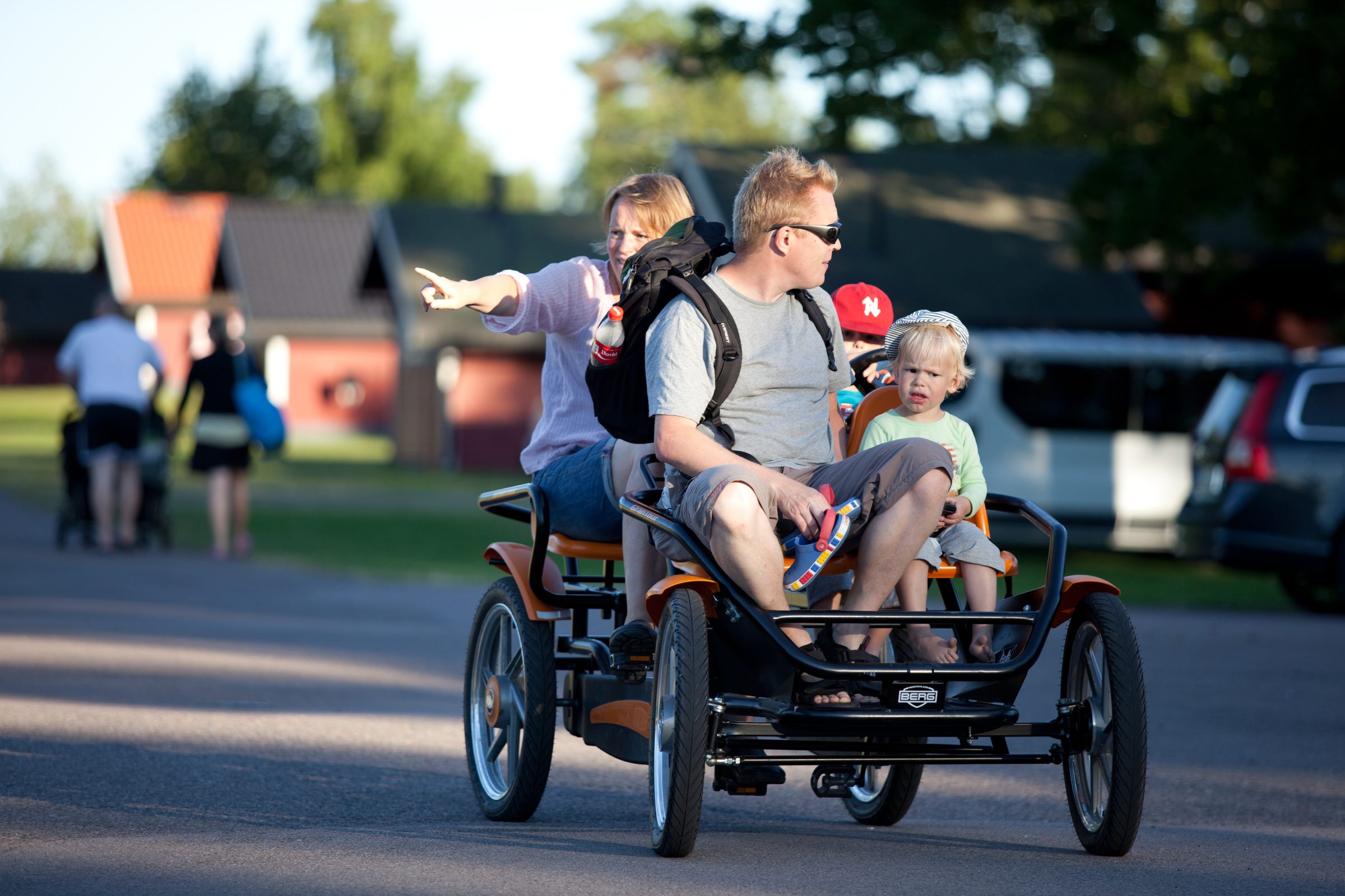 Familj som kör cykelbil på grusvägen genom området, med lekplats och stugor bakom